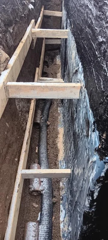 Exterior foundation waterproofing trench with drainage pipe installation and wooden bracing