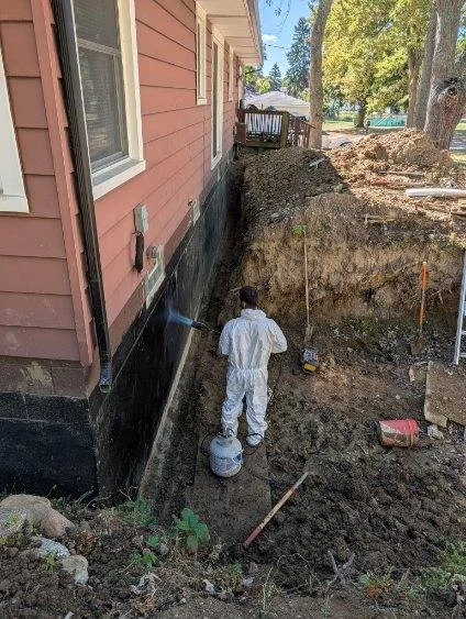 Worker in protective gear applying waterproofing treatment to exposed foundation wall during excavation