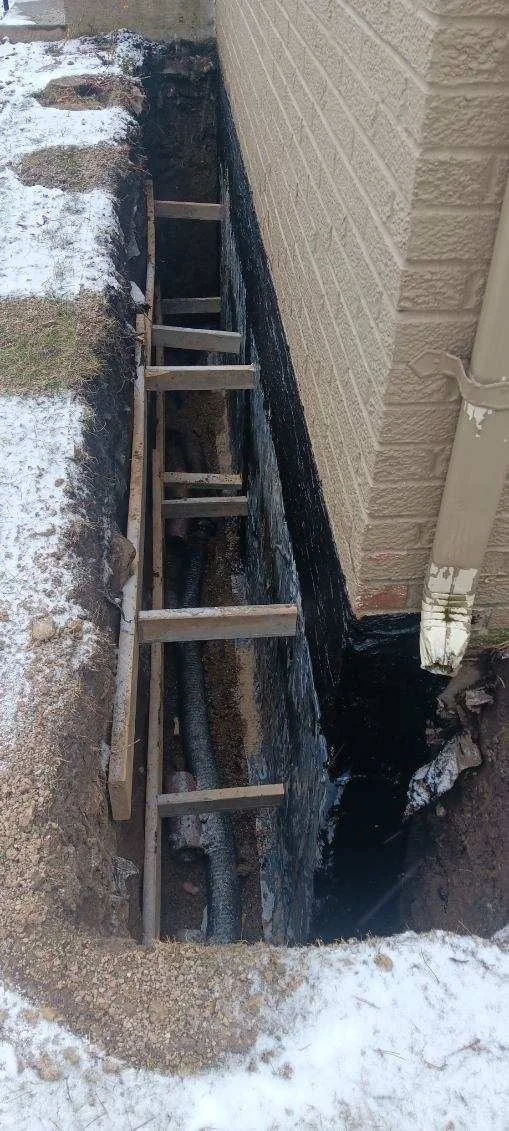 Exterior foundation wall with wooden ladder accessing below-grade waterproofing area showing moisture damage