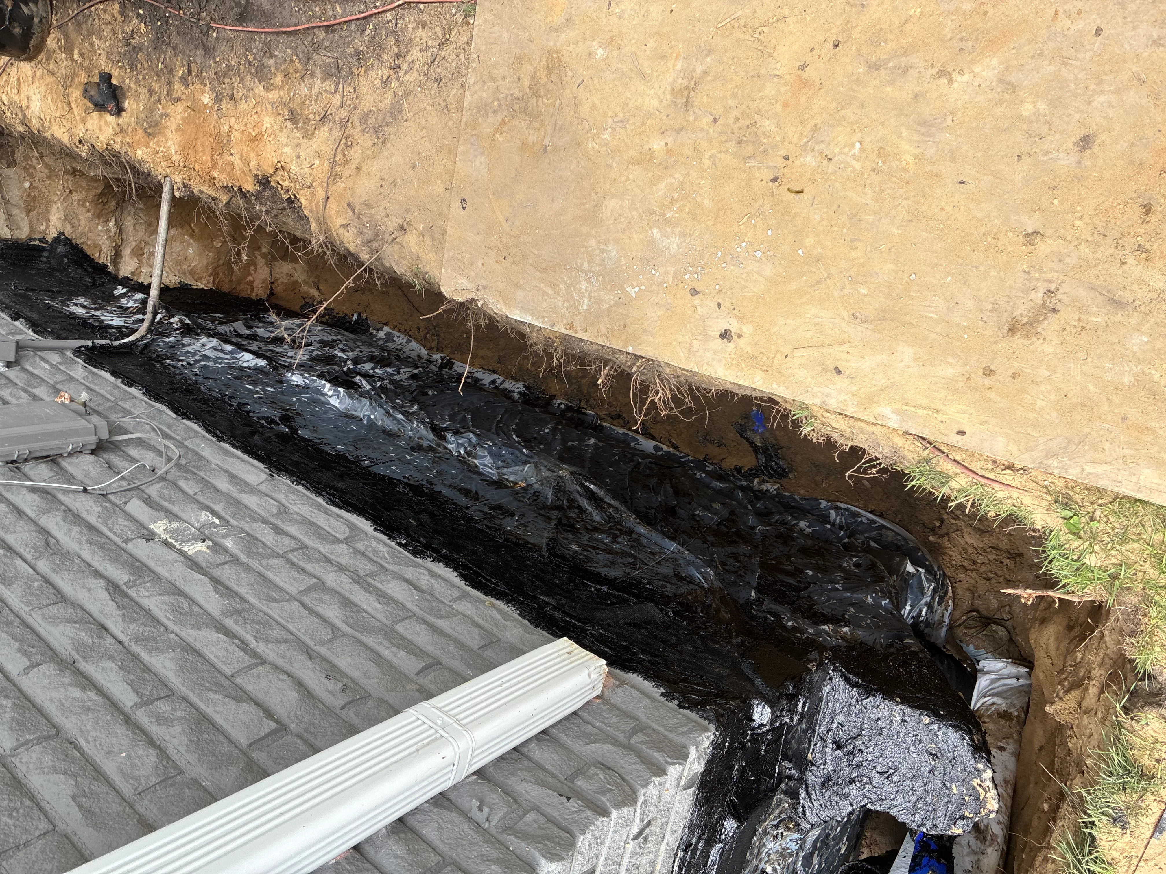 Black waterproofing membrane and white drainage pipe installed along exterior foundation wall